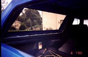 1985 Ford Bronco - The photo shows the interior of a blue vehicle, looking out through the rear window at a residential area.