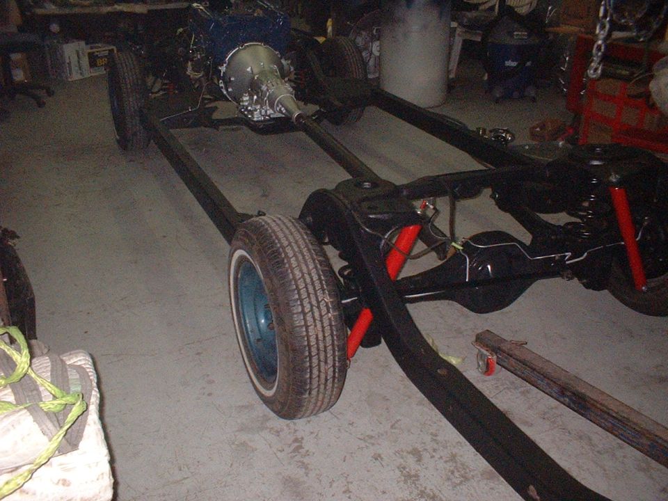 Bare car chassis frame with rear leaf-spring suspension, red shocks, transmission and blue wheel in a garage