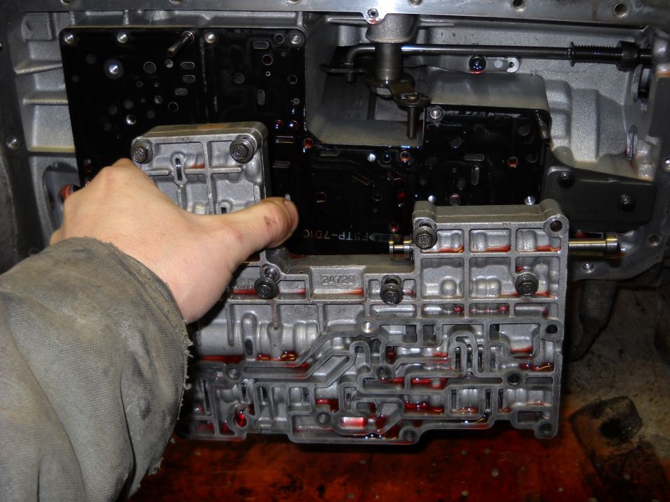 Transmission valve body with a hand holding a component, showing internal channels with red fluid