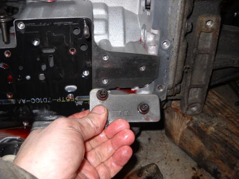 1993 Ford C-150 (Centurion Conversions) - A hand holds a metal plate with 'DC' stamped on it, near a transmission component.