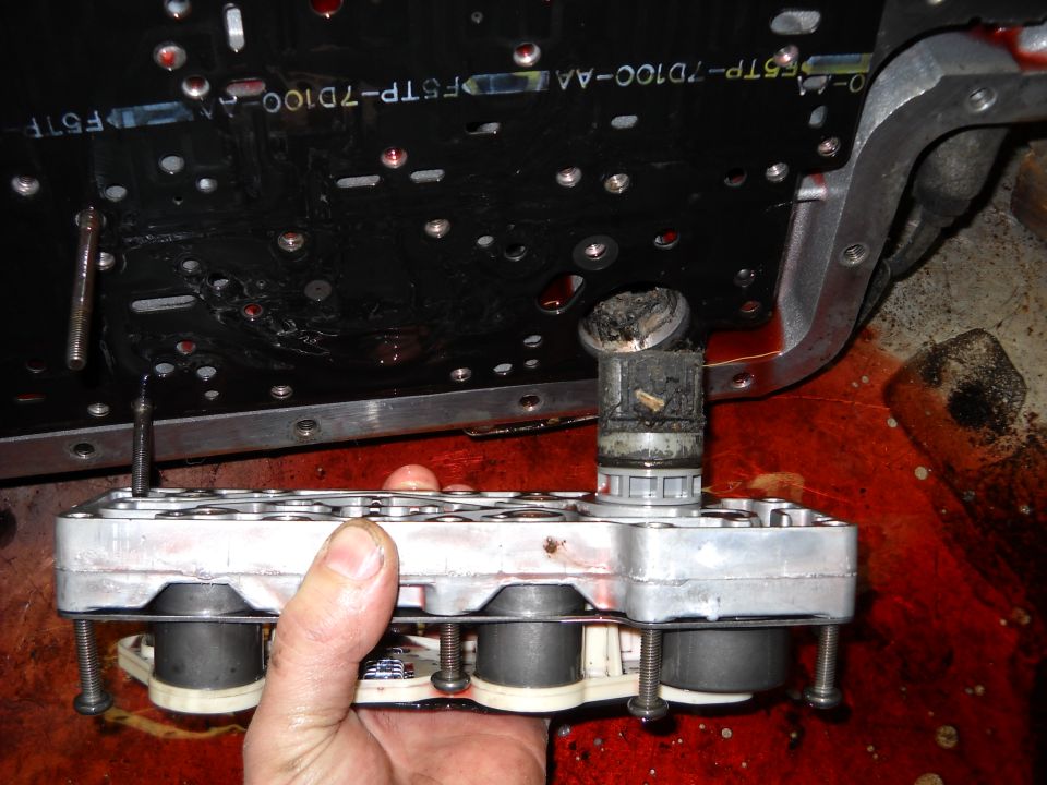 Hand holding a transmission valve body with a circuit board behind it, red fluid spilled around