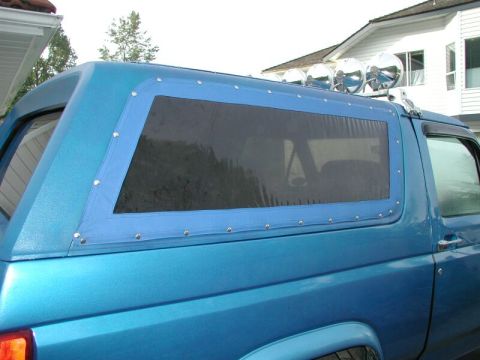 1985 Ford Bronco - Side view of a blue SUV with a custom soft top rear window and roof-mounted lights.