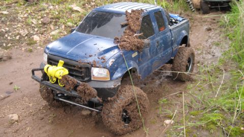 1988 Ford Bronco - A blue pickup truck is covered in mud as it navigates a wet, off-road path.