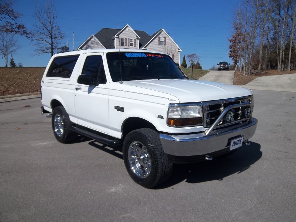 White Ford Bronco parked on a paved surface in front of houses. It has a chrome grille guard with lights and chrome wheels