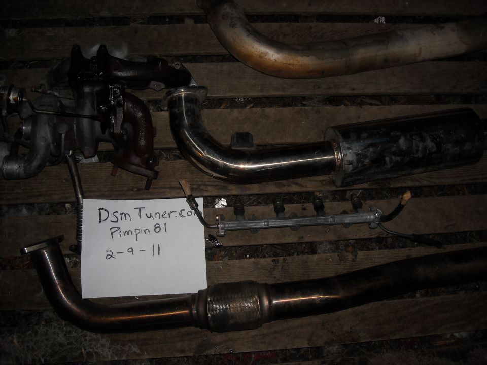 Turbo, exhaust pipe, muffler, and fuel rail laid out on wood planks. Paper note says 'Dsm Tuner.com Pimpin 81 2-9-11'