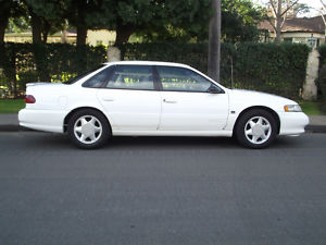 1993 Ford Taurus - A white 1993 Ford Taurus sedan is parked on the side of a street with trees and houses in the background.