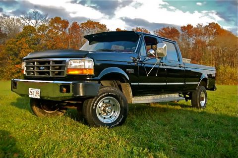 1997 Ford F-350 - This black 1997 Ford F-350 crew cab pickup truck is shown in a three-quarter front view, parked on a grassy field with autumn trees in the…