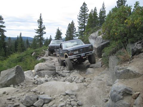 1986 Ford Bronco - Two Ford Broncos traverse a challenging rocky off-road path with a scenic mountain backdrop.