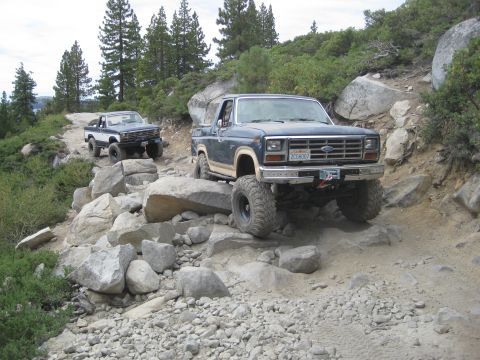 1986 Ford Bronco - Two off-road vehicles, including a Ford Bronco, traverse a challenging rocky terrain on a wooded mountainside.