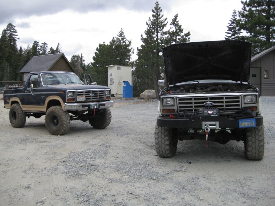 Two lifted Ford trucks parked on a gravel lot, one with its hood open