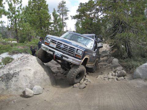 1986 Ford Bronco - Ford Bronco navigating a challenging off-road course with large boulders and trees.