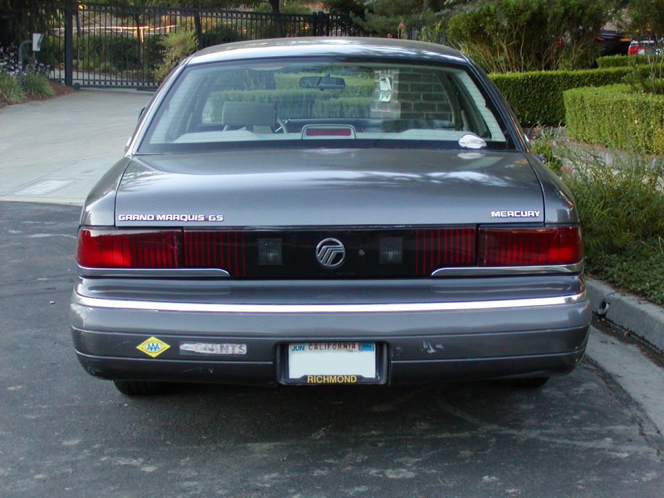 Rear view of a gray Mercury Grand Marquis GS sedan with full-width tail lamp panel, parked near hedges