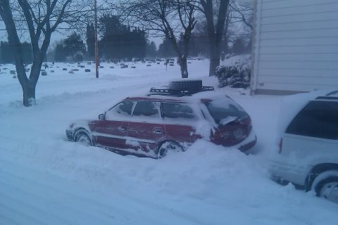 1998 Subaru Legacy - A red station wagon and a white minivan are buried in snow, with a cemetery in the background.