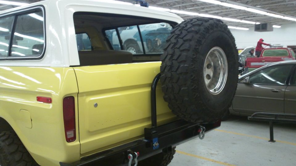 Yellow 1978 Ford Bronco rear quarter panel with spare tire mounted on swing away carrier. Other vehicles visible in background