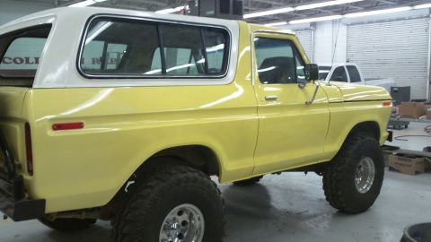 1978 Ford Bronco - A yellow 1978 Ford Bronco with a white hardtop is parked indoors, showing its lifted suspension and large off-road tires.