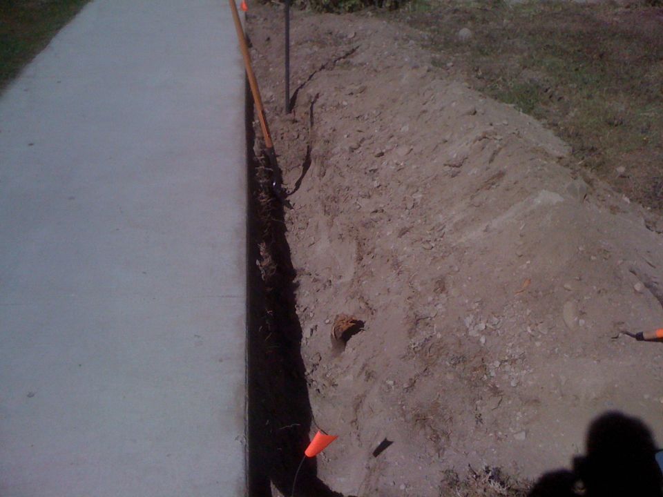 Trench dug next to a concrete sidewalk, with orange markers visible