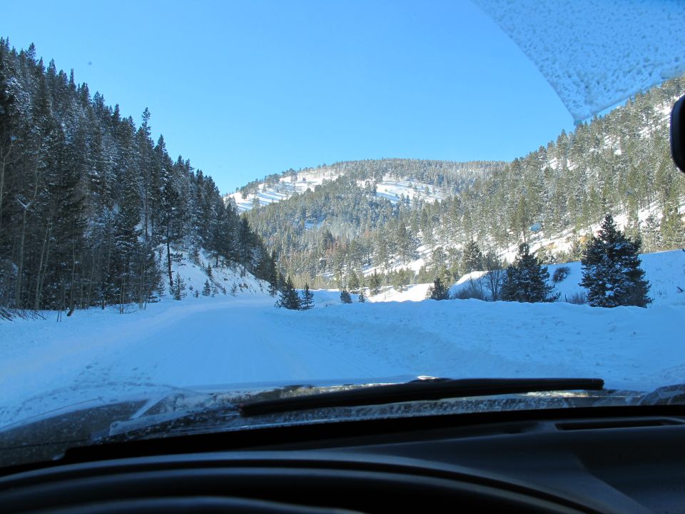 Windshield view over dark dashboard of a snowy mountain road lined with pine trees under blue sky