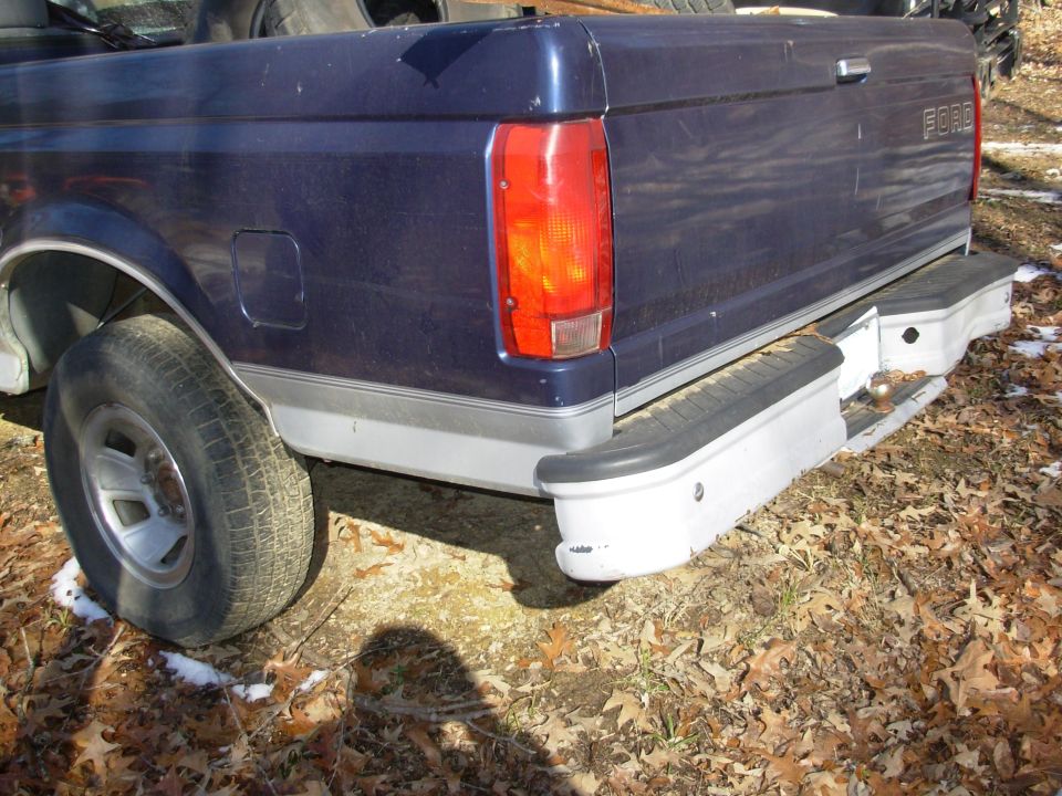 Rear quarter of dark blue Ford pickup with chrome step bumper and hitch ball, parked on leaf-covered ground