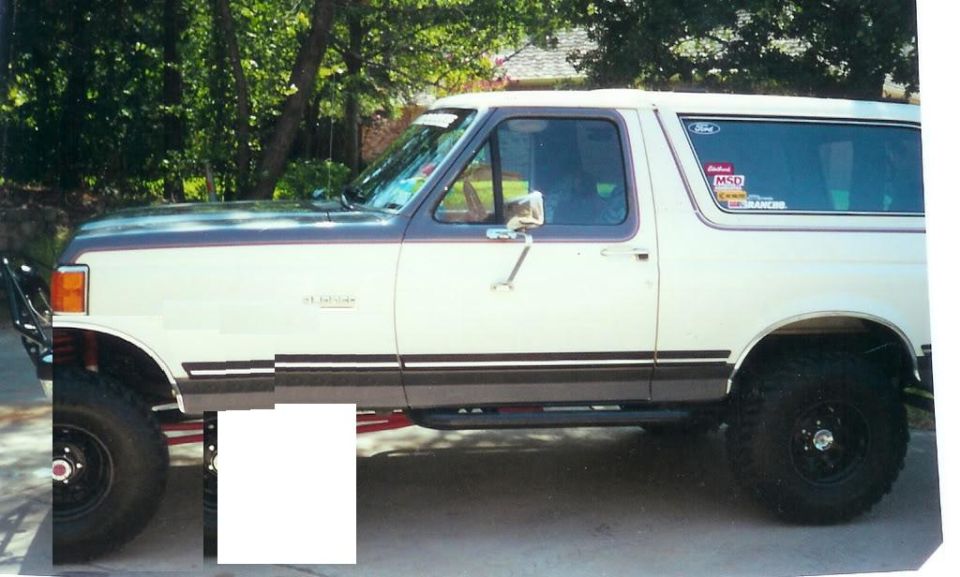 Side view of a lifted Ford Bronco with aftermarket wheels and tires. It has a two-tone paint job and some stickers on the rear window