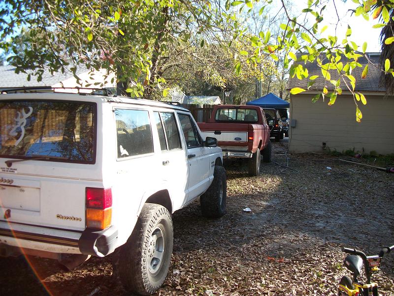 White Jeep Cherokee and red Ford pickup truck parked in a yard with a blue tarp in the background