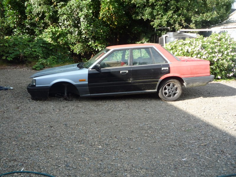 Mismatched silver, black and red 1980s sedan missing its front wheel on gravel