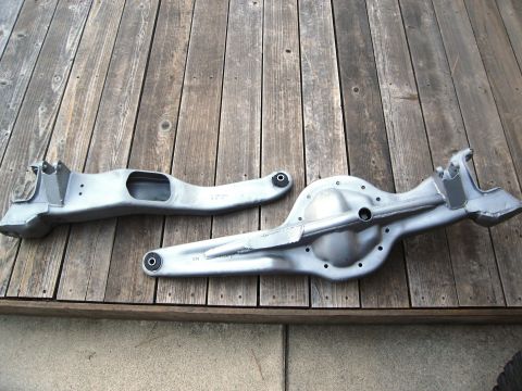 1986 Ford Bronco - Two unpainted metal automotive parts, likely from a vehicle's suspension, are laid out on a weathered wooden surface.