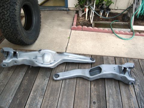 1986 Ford Bronco - Two automotive suspension components, a front axle beam and radius arms, are displayed on a wooden surface next to a tire.