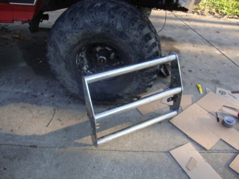 1986 Ford Bronco - A chrome grille guard lies on the ground next to a large tire of a red vehicle, with tools and cardboard scattered nearby.