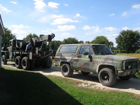 1985 Chevrolet Blazer - A camouflage-painted SUV is being towed by a large military truck on a dirt road under a blue sky.