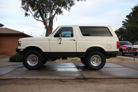 1987 Ford Bronco - A lifted white Ford Bronco is parked on a concrete driveway with a red truck in the background.