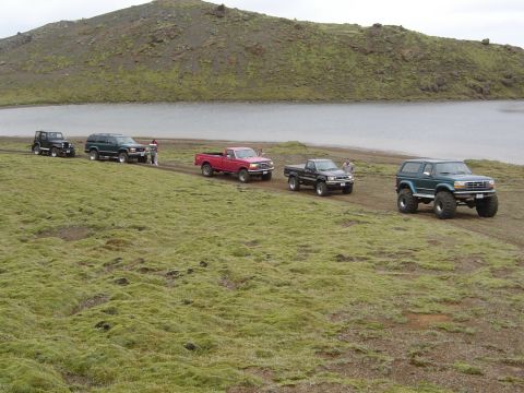 1996 Ford Bronco - Several off-road vehicles are parked on a grassy hill overlooking a lake under a cloudy sky.