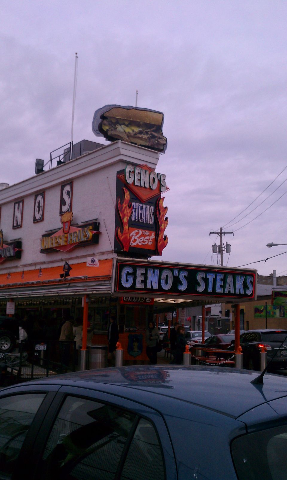 Geno's Steaks building with large sandwich sign, cars parked nearby, and a blue car in the foreground