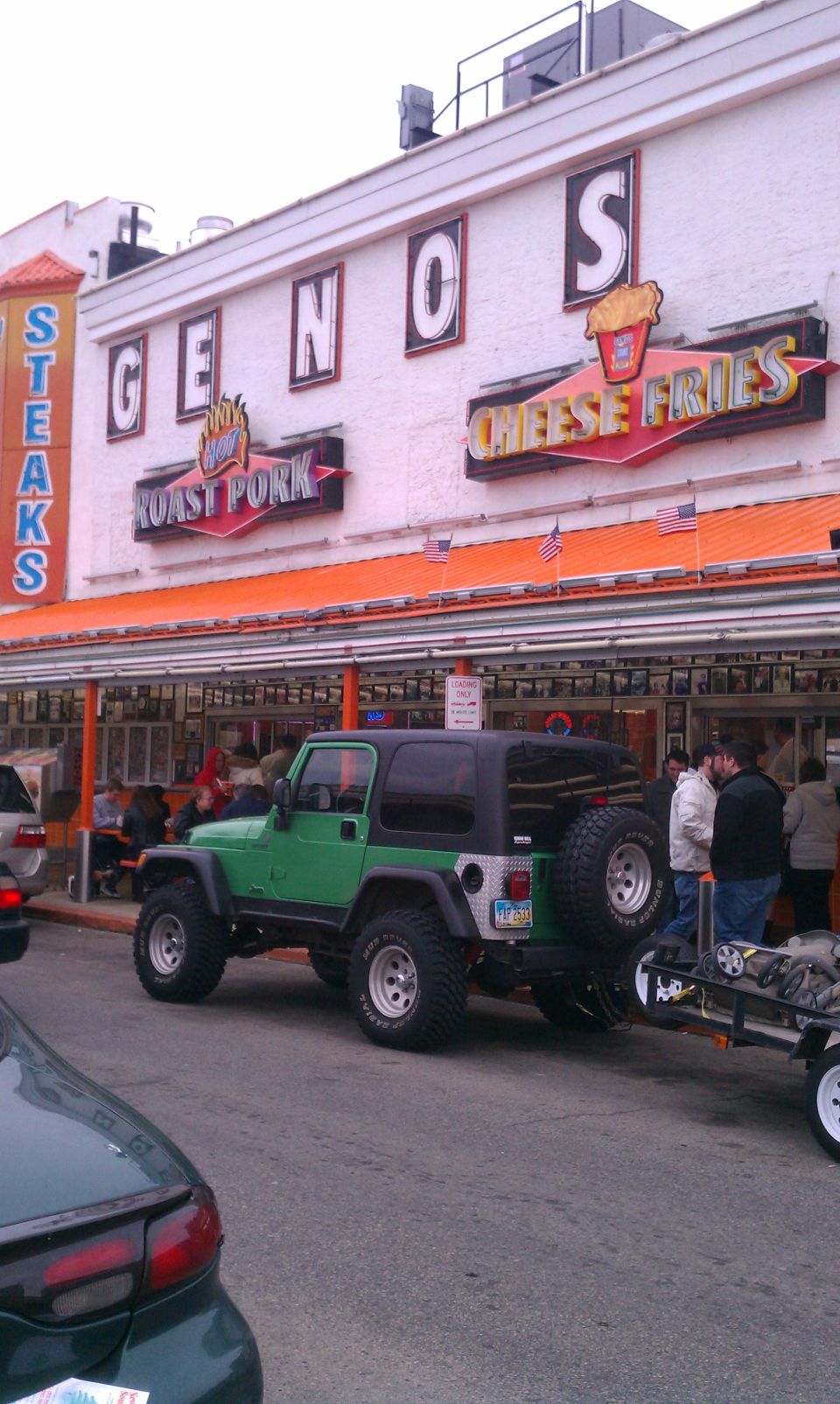 Green Jeep Wrangler with a trailer parked in front of Geno's Steaks