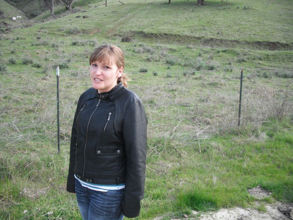 Woman in a black leather jacket and blue jeans stands in a grassy field with a fence behind her