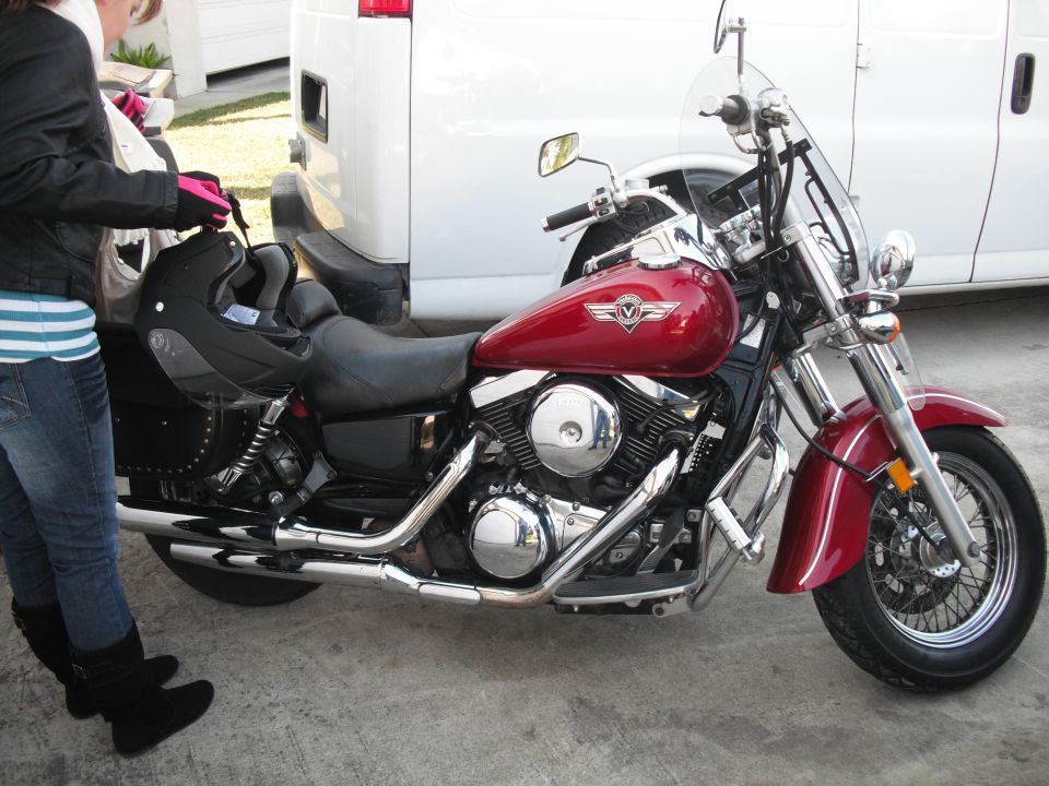 Red motorcycle with a black seat and saddlebags, parked next to a white van. A person in jeans and a black jacket is standing nearby