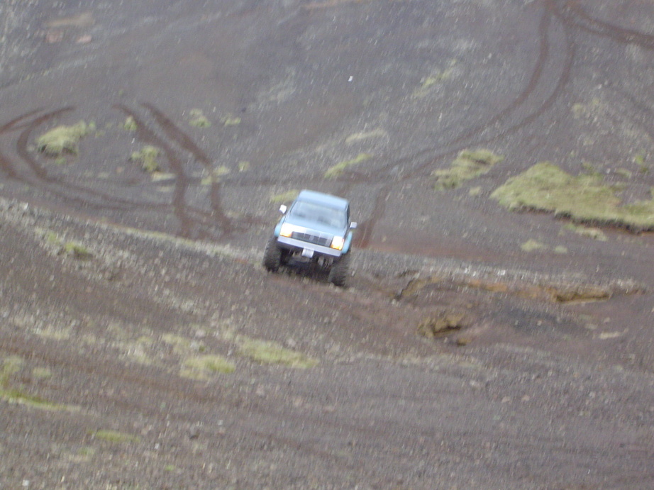 Blue 4x4 pickup truck driving across a barren gray volcanic ash slope with tire tracks