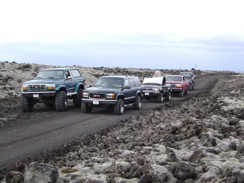 1996 Ford Bronco - A convoy of SUVs, including a lifted green 1996 Ford Bronco, travels on a rocky dirt road under an overcast sky.
