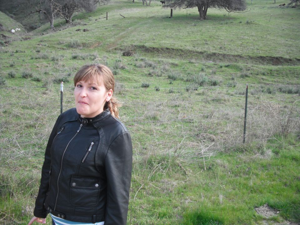 Woman in a black leather jacket stands in a grassy field with trees and a fence in the background