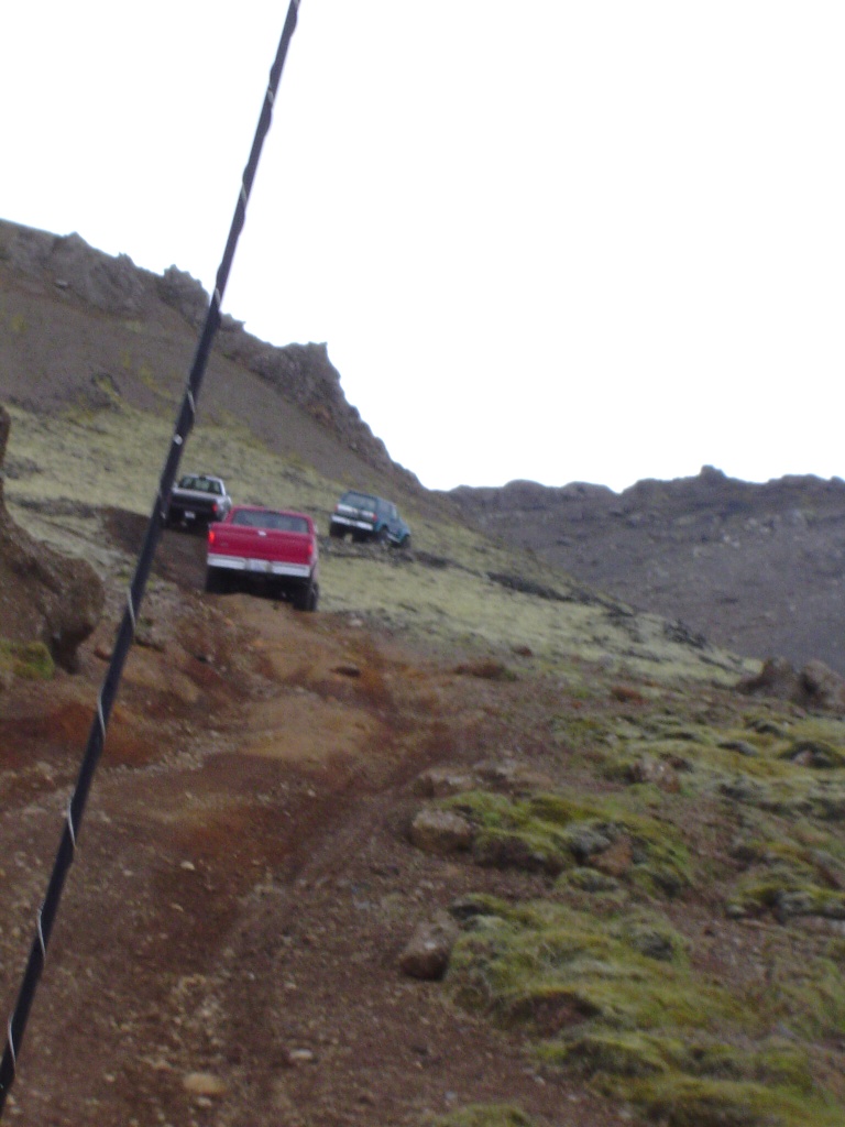 Red pickup and two SUVs parked on a rugged mossy volcanic slope under gray sky