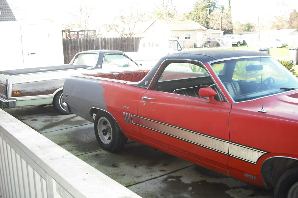 1970 Ford Ranchero in red with gray primer on the rear quarter panel, parked next to a white and woodgrain Ranchero