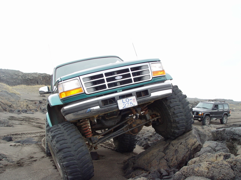 Green lifted Ford Bronco on big tires climbing dark rocks, Jeep Cherokee behind
