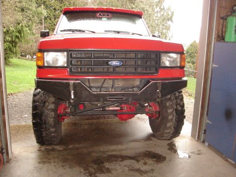 1989 Ford Bronco - Front of a lifted red Ford truck with ARB sticker on hood, custom black tubular bumper, large mud tires and red shocks, in a garage.