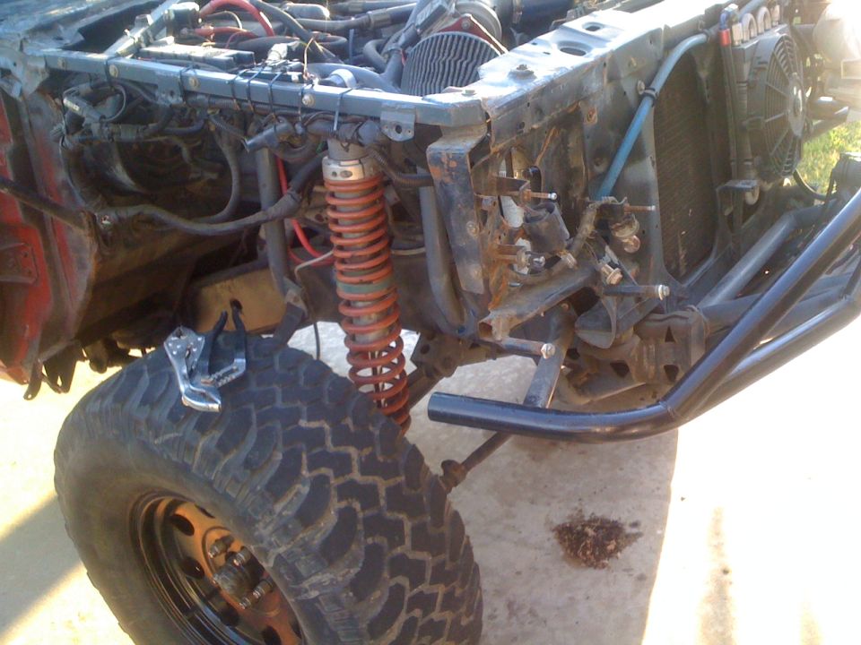 Disassembled vehicle front end with exposed engine components, red coilover shocks, and large off-road tire. A Vise-Grip is on the tire