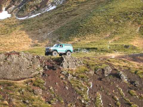 1996 Ford Bronco - A blue 1996 Ford Bronco is parked on a rocky ledge overlooking a vast, grassy, and mountainous landscape.