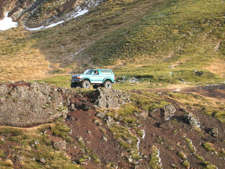 Teal and white Ford Bronco perched on a rocky outcrop high on a grassy mountain slope