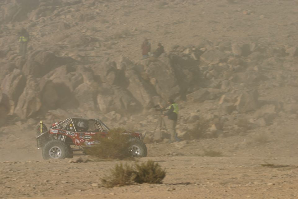 Red off-road buggy with roll cage driving on dusty desert terrain. People and camera on tripod visible in background
