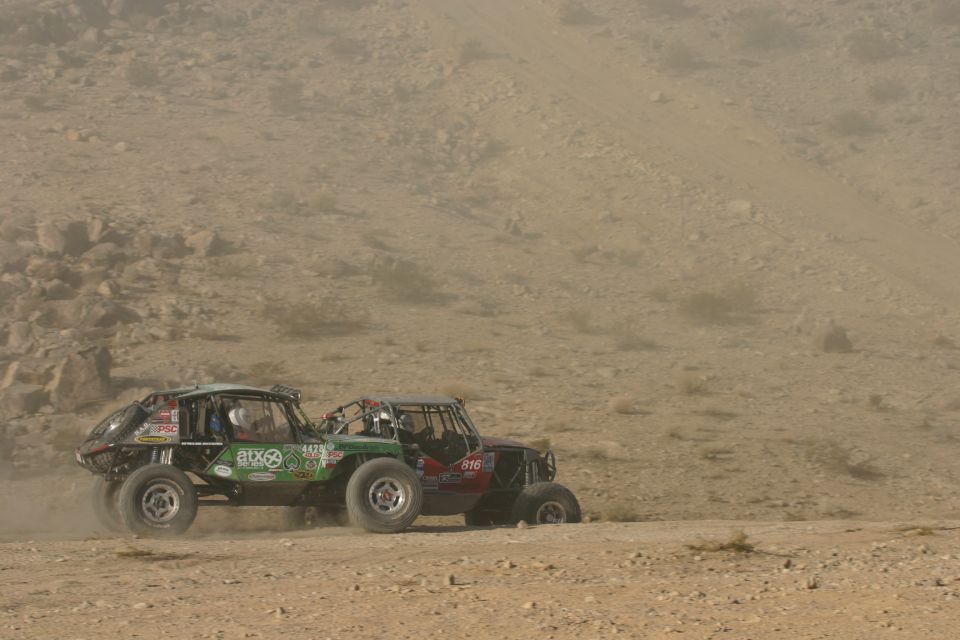 Two off-road buggies race on a dusty desert trail. One is green with sponsors, the other is red with number 816