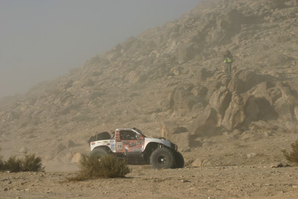 White off-road truck with large tires driving on a dusty desert terrain