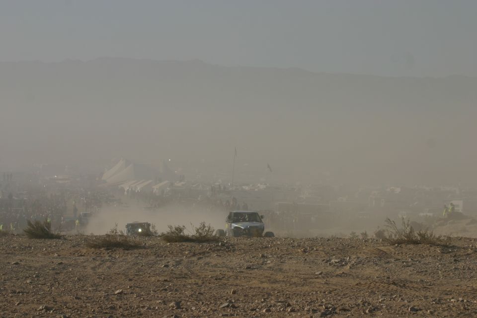 Two off-road vehicles kicking up dust on a rocky terrain with a hazy distant crowd and tents