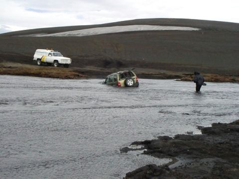 1996 Ford Bronco - A person wades through a river towards a submerged SUV, while a white truck waits on the bank.